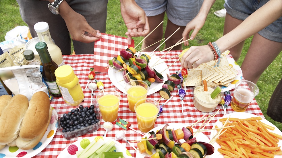 View of a picnic table with vegetable skewers, bread roles, dips, and drinks